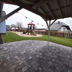 Das Bild zeigt eine Außenansicht eines Spielbereichs in einer Kindertagesstätte. Im Vordergrund liegt ein steinerner Platz mit grauen Fliesen. An einer Seite befindet sich ein Spielturm mit einer Rutsche und einem Klettergerüst. Um den Spielbereich sind grüne Rasenflächen angelegt, die eine einladende Atmosphäre schaffen. Bunte Wimpel hängen an einem Dachvorsprung und sorgen für einen fröhlichen Akzent. Im Hintergrund sind die Gebäude der Kita sowie die umliegende Nachbarschaft sichtbar, was den Bereich freundlich und einladend erscheinen lässt.