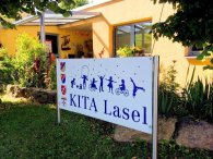 Das Bild zeigt ein Schild mit der Aufschrift „KITA Lasel“, das vor einem hellen Gebäude aufgestellt ist. Umgeben von grünem Gras und blühenden Pflanzen, vermittelt das Schild einen einladenden Eindruck. Nach oben hin sind Silhouetten von spielenden Kindern zu sehen, die Aktivitäten wie Radfahren und Tanzen darstellen. Das Gesamtbild strahlt eine fröhliche und lebendige Atmosphäre aus, passend für eine Kindertagesstätte.