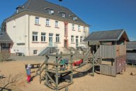 Das Bild zeigt einen Spielplatz vor dem großen, mehrstöckigen KiTa-Gebäude. Auf dem Spielplatz befindet sich eine Holz-Kletterstruktur mit Rutschen und Seilen, die den Kindern verschiedene Spielmöglichkeiten bietet. Zwei Kinder spielen aktiv, eines klettert und das andere befindet sich in der Nähe der Struktur. Der Boden ist mit Sand bedeckt, und der klare blaue Himmel deutet auf einen sonnigen Tag hin, was eine fröhliche und einladende Atmosphäre für Aktivitäten im Freien schafft.
