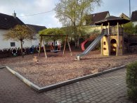 Der Spielplatz bietet eine vielfältige Outdoor-Spielumgebung für Kinder. In der Mitte steht ein Spielturm mit einer Rutsche und einem Kletterbereich. Auf der linken Seite befindet sich eine Schaukel. Der Boden ist mit Holzspänen bedeckt, was eine sichere Landefläche bietet. Außerdem sind die umgebenden Wände aus Holz, die eine geschützte Atmosphäre schaffen. Grüne Bäume und Büsche sorgen für einen naturbelassenen Look und Schatten, wodurch der Spielplatz einladend und abenteuerlich wirkt.
