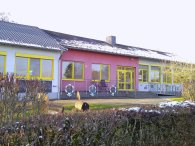 Das Gebäude ider Kindertagesstätte Weinsheim st eine einzelgeschossige Einrichtung. Die Fassade ist in hellen Farben gestaltet, mit einem pinkfarbenen Teil und weißen Wänden, die von leuchtend gelben Fensterrahmen umgeben sind. Große Fenster sorgen für ausreichend Licht und einen einladenden Blick nach außen. Vor dem Gebäude sind Blumenillustrationen und gestaltete Geländer sichtbar, die die kinderfreundliche Atmosphäre unterstreichen.