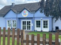 Das Bild zeigt das Eingangsgebäude der Kita Winterspelt. Die Fassade ist in einem freundlichen blauen Farbton gehalten, und über der Eingangstür befindet sich ein buntes Schild mit dem Schriftzug "KITA". Vor dem Gebäude steht ein Holzlattenzaun, der den Eingang umgibt. Die Fenster des Hauses sind groß, was einen hellen und einladenden Eindruck vermittelt. Im Hintergrund sind einige Grünflächen und Bäume zu sehen, die eine angenehme Umgebung schaffen.